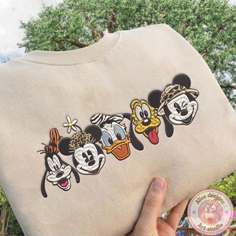 Puede incluir: Sudadera beige con caras bordadas de personajes de dibujos animados: Goofy, Mickey Mouse, Pato Donald, Pluto y Mickey Mouse con sombrero de safari. El dise&ntilde;o incluye una peque&ntilde;a flor y un detalle de estampado de leopardo. La sudadera es sostenida por una mano.
