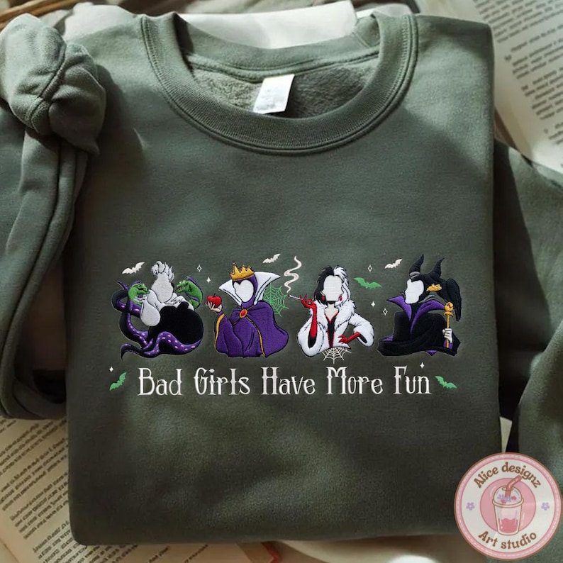 May include: An olive green sweatshirt featuring embroidered cartoon villains. The design includes Ursula, the Evil Queen, Cruella de Vil, and Maleficent. The text "Bad Girls Have More Fun" is also embroidered on the sweatshirt.