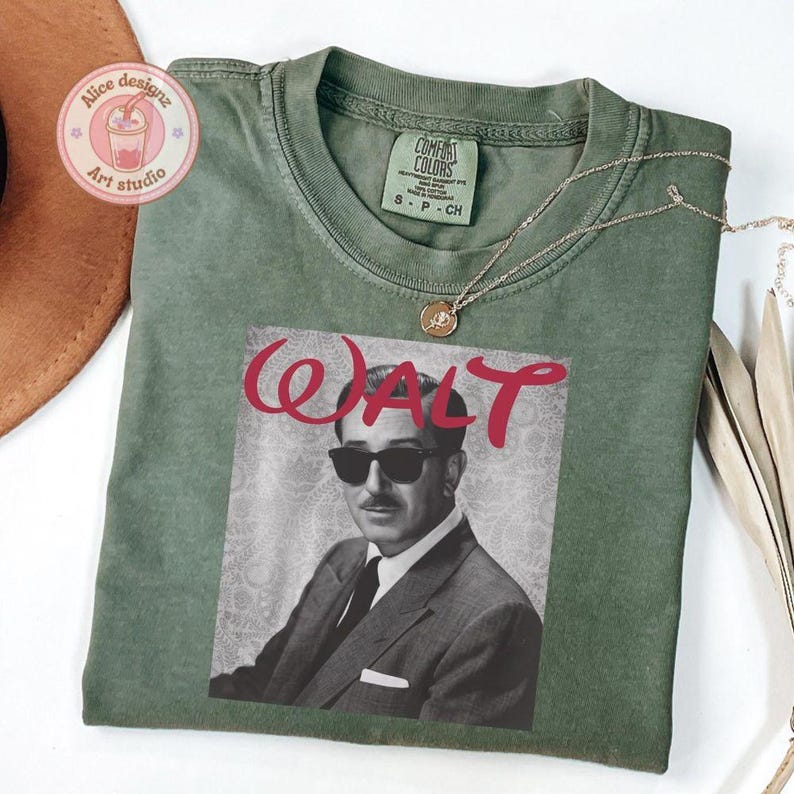 Op de afbeelding: Een olijfgroen t-shirt met een zwart-wit afbeelding van Walt Disney met een zonnebril en de naam "WALT" in rood script. Een gouden ketting met een hanger is over het shirt gedrapeerd. Het shirt is van het merk Comfort Colors, maat S-P-CH.