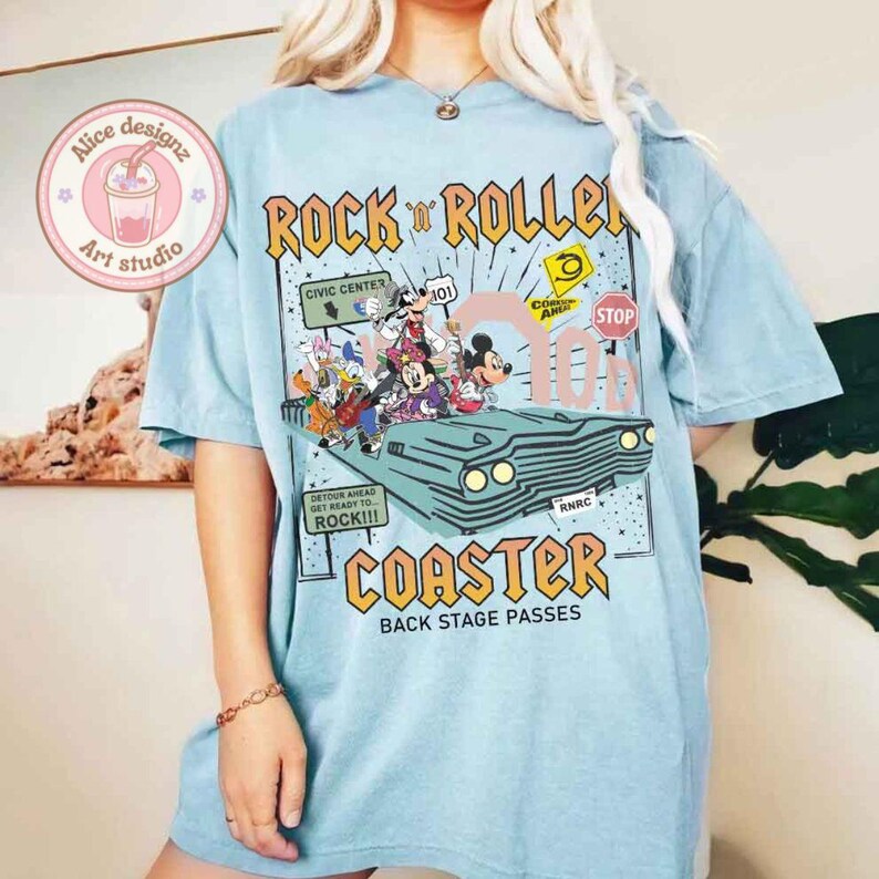 May include: A light blue t-shirt with a graphic print of Mickey Mouse and other Disney characters riding in a vintage car. The text "Rock 'n' Roller Coaster" is printed in a bold, black font with yellow accents. The text "Back Stage Passes" is printed in a smaller font below the graphic.