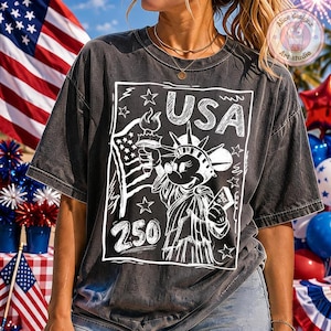May include: Dark gray t-shirt featuring a white graphic of the Statue of Liberty with Mickey Mouse's face, holding a torch and a scroll that says "July". The word "USA" is above the image, and the number "250" is below. The shirt has a vintage, faded look.