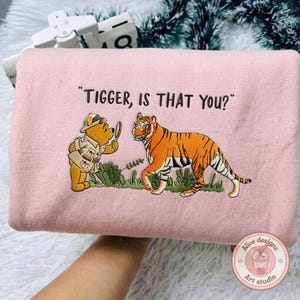 Op de afbeelding: Zachtroze sweatshirt met een geborduurd ontwerp van Winnie de Poeh en een tijger, met de tekst "TIGGER, IS THAT YOU?". Het ontwerp toont een cartoonbeer met een vergrootglas. De sweatshirt heeft een ronde hals.