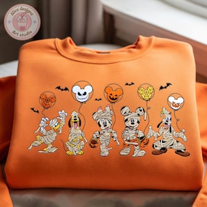 May include: Orange sweatshirt featuring a Halloween-themed design. The design includes cartoon characters dressed as mummies, holding jack-o'-lanterns, and balloons with spiderwebs and spooky faces. Bats are also included in the design.