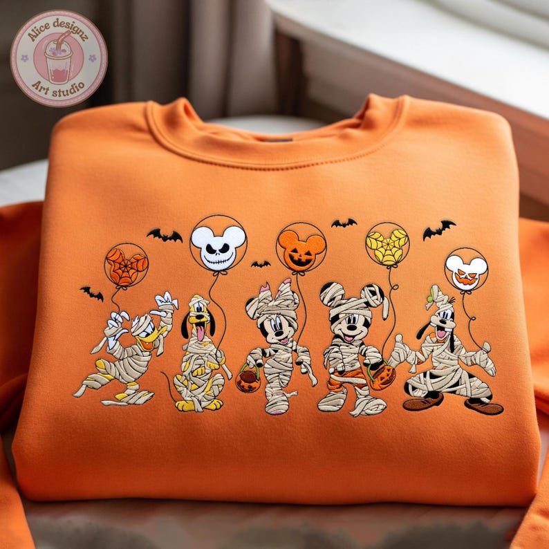 May include: Orange sweatshirt featuring a Halloween-themed design. The design includes cartoon characters dressed as mummies, holding jack-o'-lanterns, and balloons with spiderwebs and spooky faces. Bats are also included in the design.