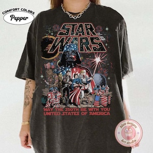 Op de afbeelding: Donkergrijs t-shirt met een Star Wars-ontwerp met Darth Vader, vuurwerk en Amerikaanse vlaggen. De tekst luidt "MAY THE 250TH BE WITH YOU UNITED STATES OF AMERICA". Het shirt heeft een Comfort Colors-label.