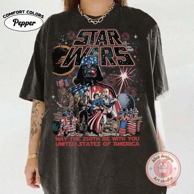 Op de afbeelding: Donkergrijs t-shirt met een Star Wars-ontwerp met Darth Vader, vuurwerk en Amerikaanse vlaggen. De tekst luidt "MAY THE 250TH BE WITH YOU UNITED STATES OF AMERICA". Het shirt heeft een Comfort Colors-label.