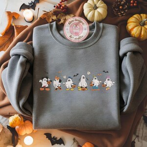 K&ouml;nnte beinhalten: Ein graues Sweatshirt mit einem Halloween-Motiv. Das Design zeigt Cartoon-Geister, die als beliebte Charaktere verkleidet sind, K&uuml;rbisse halten und von Flederm&auml;usen begleitet werden. Das Sweatshirt hat lange &Auml;rmel und einen Rundhalsausschnitt.