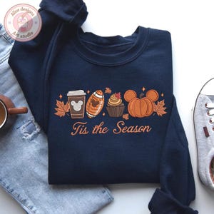 May include: Navy blue sweatshirt with an embroidered fall-themed design. The design includes a coffee cup, football, cupcake, and pumpkin, with the text "Tis the Season" in orange. The sweatshirt is paired with blue jeans and a sneaker.
