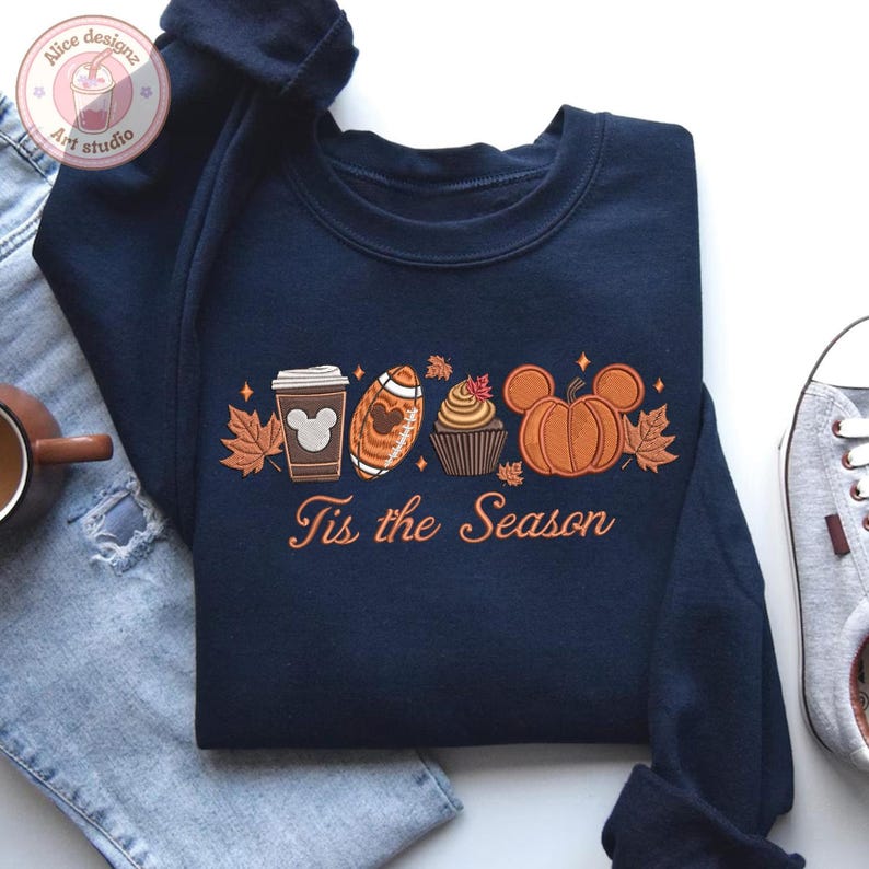 May include: Navy blue sweatshirt with an embroidered fall-themed design. The design includes a coffee cup, football, cupcake, and pumpkin, with the text "Tis the Season" in orange. The sweatshirt is paired with blue jeans and a sneaker.