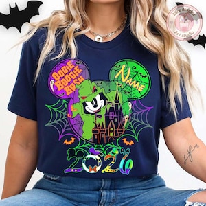 May include: Navy blue t-shirt featuring a Halloween-themed design. The graphic includes a cartoon character in a green costume, a haunted castle, spiderwebs, and the text "Oogie Boogie Bash" and "2026". The design is in a Mickey Mouse head shape.