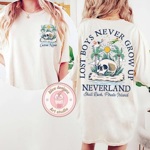 May include: A white t-shirt with a graphic print of a skull and palm trees. The text "Lost Boys Never Grow Up" is printed in blue on the back of the shirt. The text "Neverland Skull Rock, Pirate Island" is printed below the graphic.