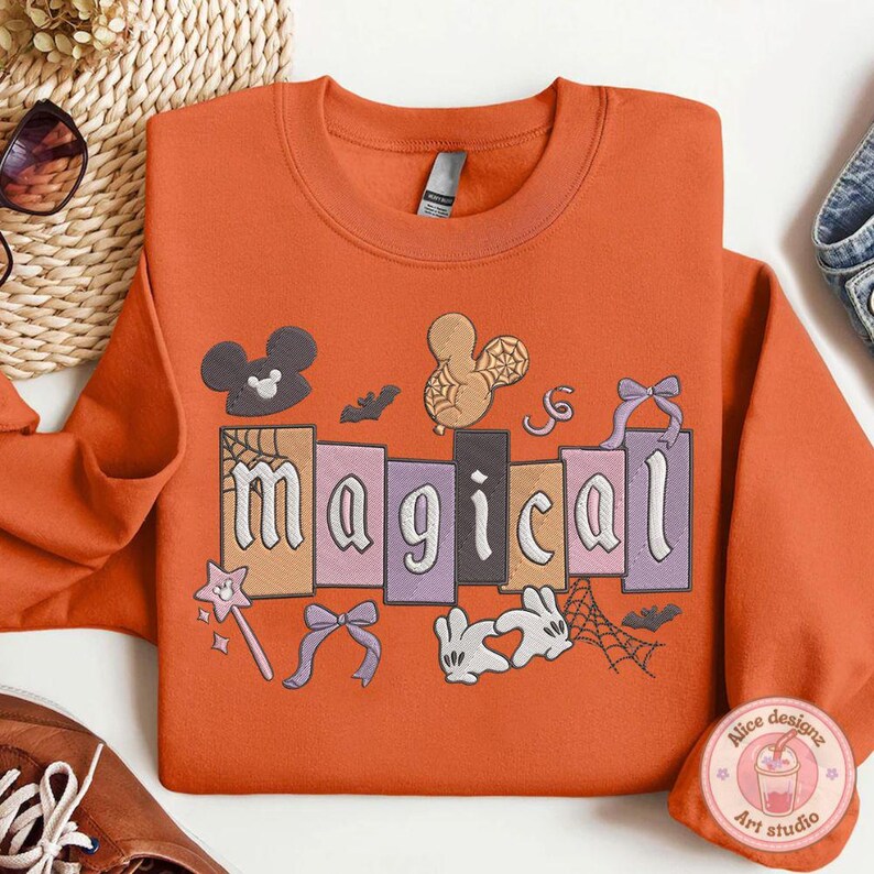 May include: Orange crew neck sweatshirt with the word "magical" in a block letter design. The sweatshirt features embroidered images of Mickey Mouse, a spider web, a magic wand, bows, and a rabbit. The sweatshirt is orange.