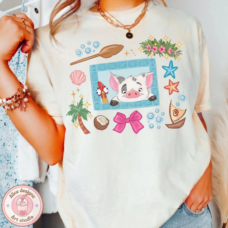 Puede incluir: Camiseta color crema con una ilustraci&oacute;n de dibujos animados de un cerdo y un gallo en un marco azul, rodeada de elementos tropicales. La camiseta tiene cuello redondo y mangas cortas. El dise&ntilde;o incluye una palmera, un lazo y un barco.