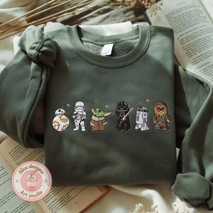 May include: Olive green sweatshirt featuring embroidered Star Wars characters: BB-8, a Stormtrooper, Grogu, Darth Vader, R2-D2, and Chewbacca. The sweatshirt has a crew neck and long sleeves. The design includes small gold stars.