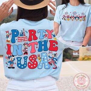Peut inclure: T-shirt bleu clair avec l'inscription "Party in the USA" en rouge, blanc et bleu. Le t-shirt pr&eacute;sente des personnages de dessins anim&eacute;s et des images patriotiques, notamment des feux d'artifice et le drapeau am&eacute;ricain. Un deuxi&egrave;me t-shirt est visible avec un design similaire.