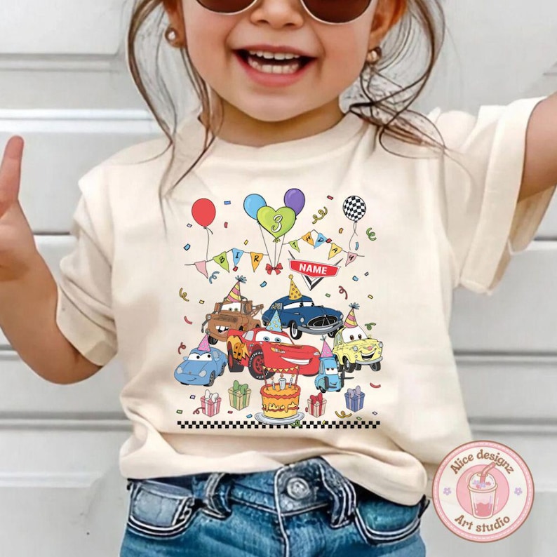 Op de afbeelding: Cr&egrave;mekleurig T-shirt met een kleurrijk cartoon auto-ontwerp met een verjaardagsthema. De afbeelding bevat verschillende auto's, ballonnen, een taart en het nummer 3. Het shirt heeft een zwart-witte geblokte rand aan de onderkant.