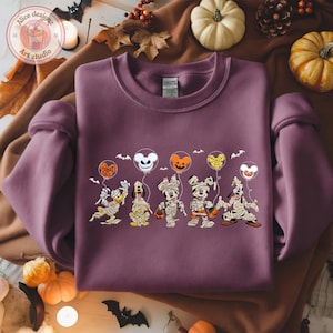 May include: A mauve sweatshirt featuring a Halloween-themed graphic. The design includes cartoon characters dressed as mummies, holding balloon-shaped pumpkins and bats. The Alice Design Art Studio logo is visible in the upper left corner.