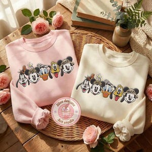 Puede incluir: Dos sudaderas, una rosa claro y otra crema, cada una con un dise&ntilde;o bordado de personajes de dibujos animados. El dise&ntilde;o incluye Mickey Mouse, el Pato Donald, Goofy y otros. Las sudaderas se exhiben sobre una superficie de madera con detalles florales.