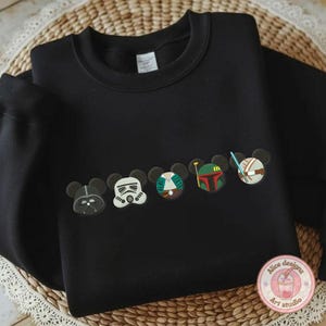 May include: Black sweatshirt featuring embroidered character designs. The designs include Darth Vader, a Stormtrooper, and other characters. The sweatshirt has a crew neck and long sleeves. The design is in the style of Mickey Mouse ears.