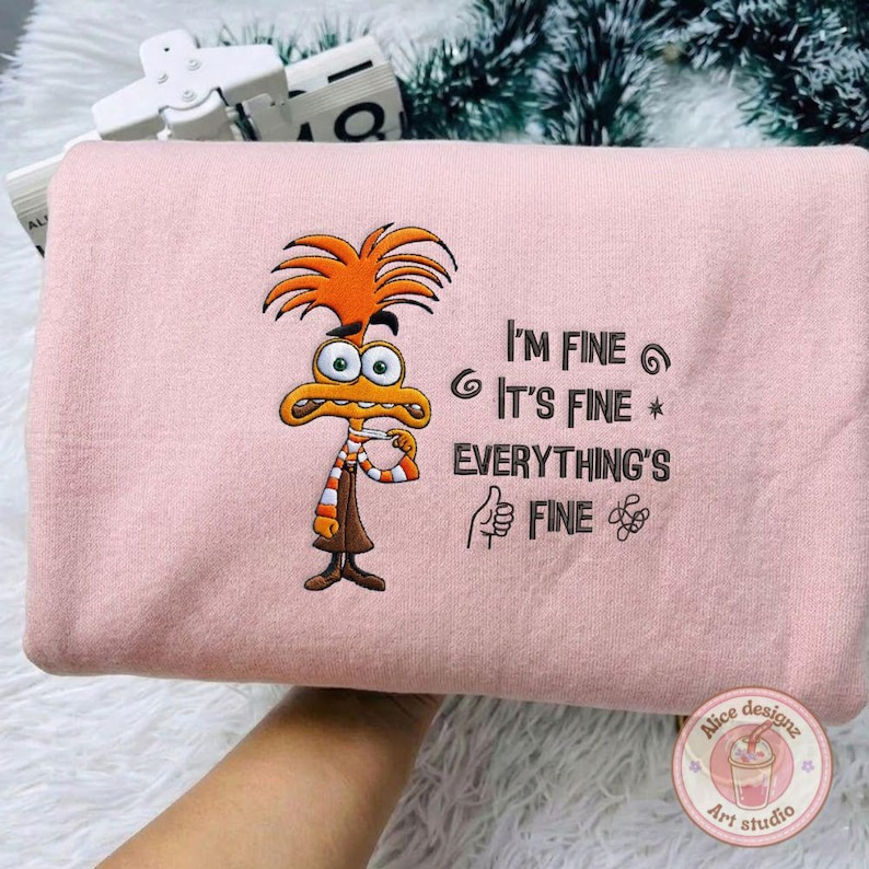 May include: A light pink sweatshirt featuring an embroidered cartoon character with orange hair and a striped scarf. The text "I'M FINE, IT'S FINE, EVERYTHING'S FINE" is also embroidered on the sweatshirt.