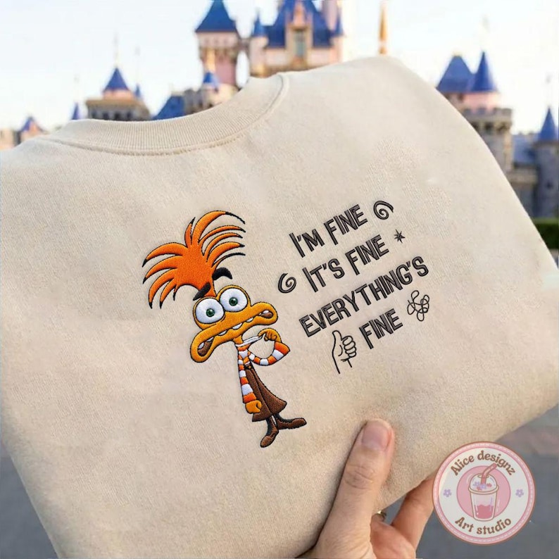 May include: Beige sweatshirt featuring an embroidered cartoon character with orange hair and a brown outfit. The text "I'M FINE, IT'S FINE, EVERYTHING'S FINE" is printed on the sweatshirt.