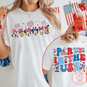 Peut inclure: T-shirt blanc avec un motif color&eacute; de personnages Disney portant des chapeaux patriotiques et des feux d'artifice. Le dos du t-shirt affiche le texte "Party in the USA" avec des illustrations de personnages Disney. Un design festif.