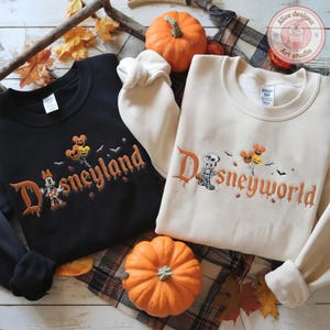 May include: A black sweatshirt with the embroidered text Disneyland and a white sweatshirt with the embroidered text Disneyworld. Both feature a skeleton character, pumpkin balloons, and bats. The text is in an orange, dripping font.