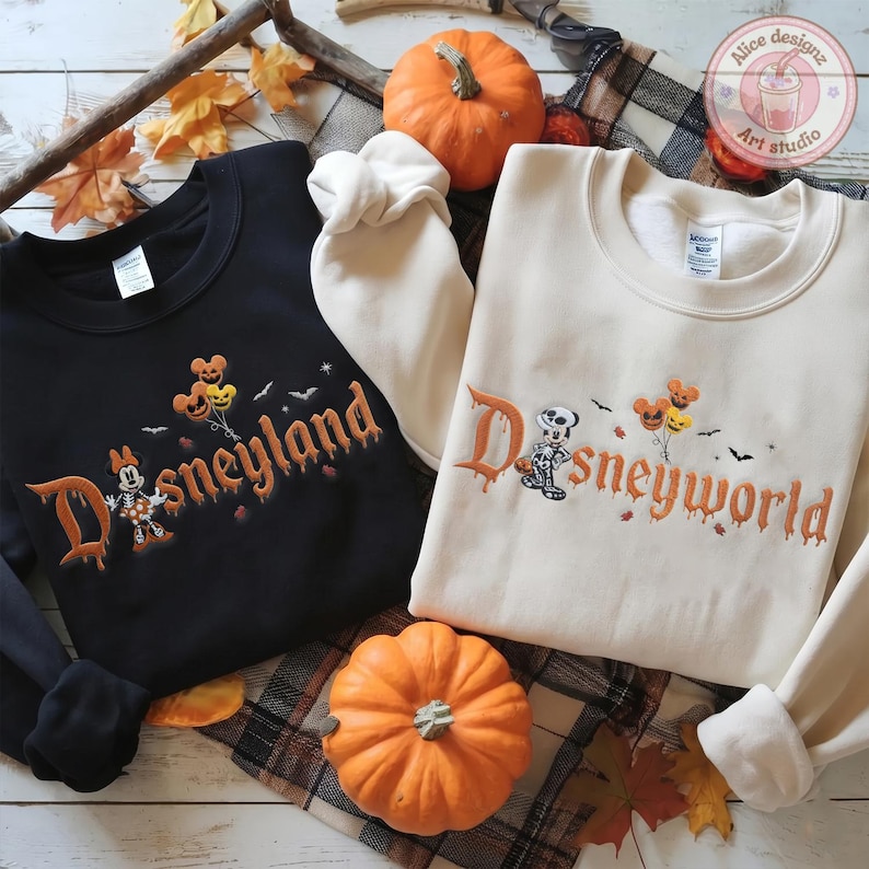 May include: A black sweatshirt with the embroidered text Disneyland and a white sweatshirt with the embroidered text Disneyworld. Both feature a skeleton character, pumpkin balloons, and bats. The text is in an orange, dripping font.