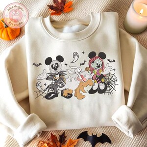 May include: Cream-colored sweatshirt featuring a Halloween-themed graphic. The design includes cartoon characters, bats, spiderwebs, and ghosts. The sweatshirt has long sleeves and a crew neck.