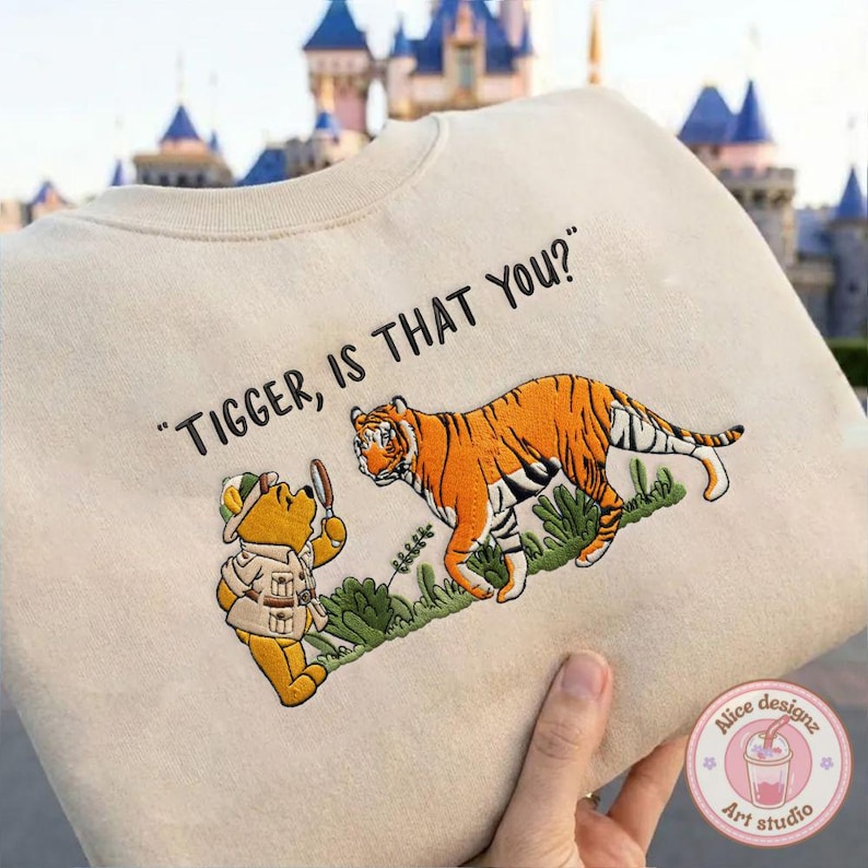Op de afbeelding: Beige sweatshirt met een geborduurd ontwerp van Winnie de Poeh en Tijger. De tekst "Tigger, is that you?" staat boven de personages. Het ontwerp toont groen gebladerte. Op de achtergrond is een kasteel te zien.