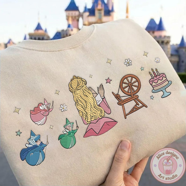 May include: Beige sweatshirt featuring an embroidered design of a princess with long blonde hair, three fairies, a spinning wheel, and a cake. The design includes stars and flowers. The sweatshirt is held by a hand.