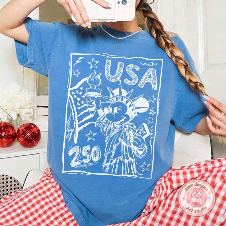 May include: A blue t-shirt with a white graphic featuring the Statue of Liberty as Mickey Mouse, the word "USA", stars, and the number "250". The American flag is also included in the design.