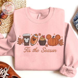May include: A light pink sweatshirt with an embroidered fall-themed design. The design includes a coffee cup, football, cupcake, and pumpkin, each with a Mickey Mouse silhouette. The text "Tis the Season" is below the design.