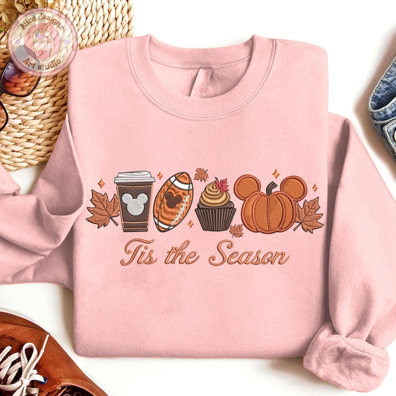 May include: A light pink sweatshirt with an embroidered fall-themed design. The design includes a coffee cup, football, cupcake, and pumpkin, each with a Mickey Mouse silhouette. The text "Tis the Season" is below the design.