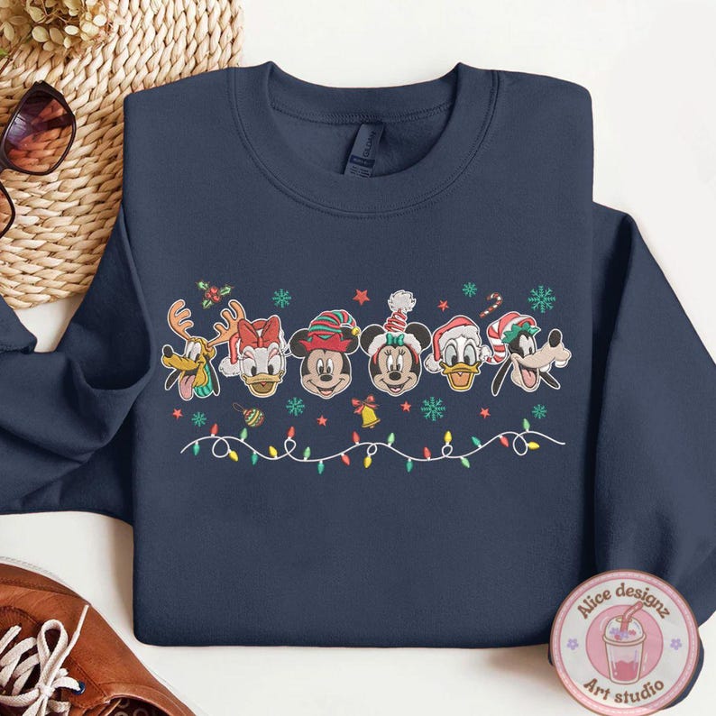 Op de afbeelding: Marineblauwe sweatshirt met een feestelijk ontwerp met cartoongezichten met kerstmutsen. Het ontwerp omvat rendieren, Donald Duck, Daisy Duck, Mickey Mouse, Minnie Mouse en Goofy, samen met kerstverlichting en sneeuwvlokken.