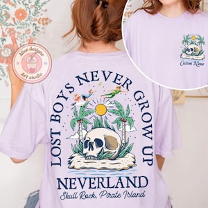May include: Purple t-shirt with a graphic print of a skull, palm trees, and flying figures. The text "Lost Boys Never Grow Up" is printed in a circle around the skull. The text "Neverland Skull Rock, Pirate Island" is printed below the graphic.