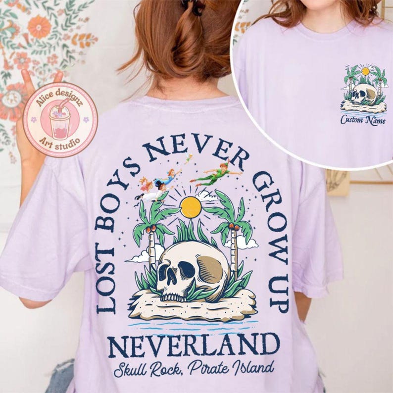 May include: Purple t-shirt with a graphic print of a skull, palm trees, and flying figures. The text "Lost Boys Never Grow Up" is printed in a circle around the skull. The text "Neverland Skull Rock, Pirate Island" is printed below the graphic.