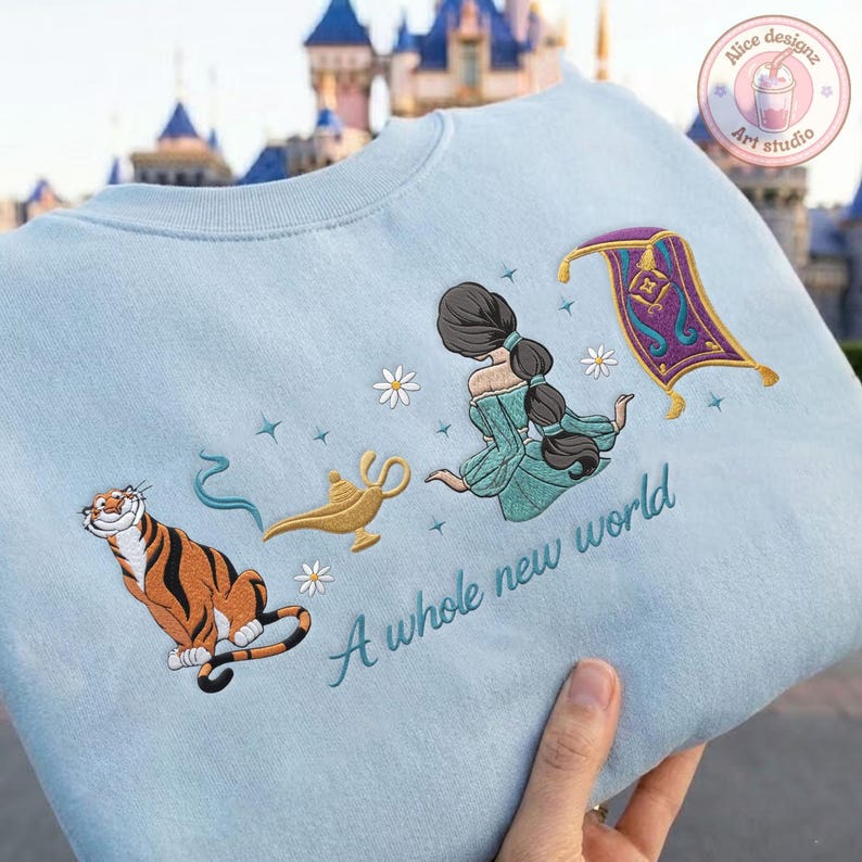May include: A light blue sweatshirt featuring embroidered designs of a tiger, a gold lamp, a character with long dark hair in a teal outfit, and a purple flying carpet. The text A whole new world is embroidered in teal script below the designs.