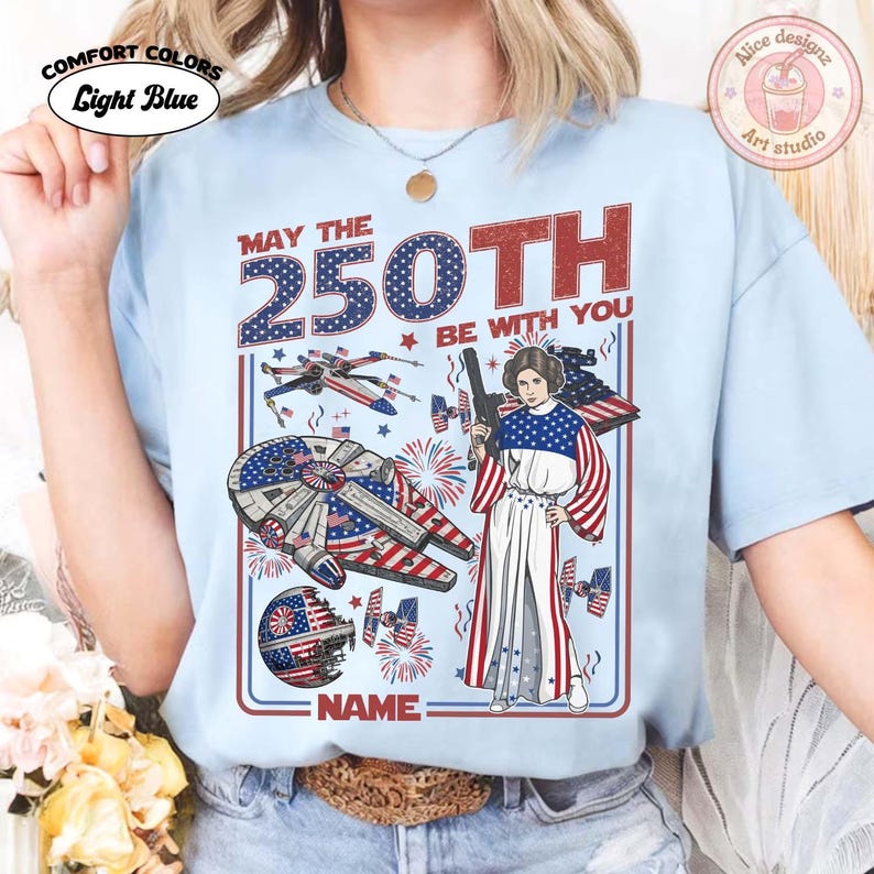 May include: Pale blue t-shirt featuring a patriotic design with the text "MAY THE 250TH BE WITH YOU". The graphic includes a spaceship, a character with a blaster, and fireworks. The shirt is labelled "COMFORT COLORS".