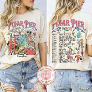 May include: A beige t-shirt with a colorful graphic of Pixar characters and the text "Pixar Pier Tour" and "Disneyland". The graphic includes characters from Toy Story, Monsters Inc, Finding Nemo, The Incredibles, Cars, Ratatouille, Wall-E, Up, Brave, Inside Out, Finding Dory, Coco, Luca, Turning Red, and Elemental. The text "Pixar Pier Tour" is in a large, bold font and the text "Disneyland" is in a smaller font.