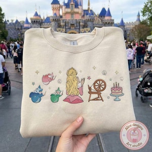 May include: Beige sweatshirt featuring a colorful graphic of a princess, fairies, a spinning wheel, and a cake. The design includes stars and flowers. The sweatshirt is held up in front of a castle.