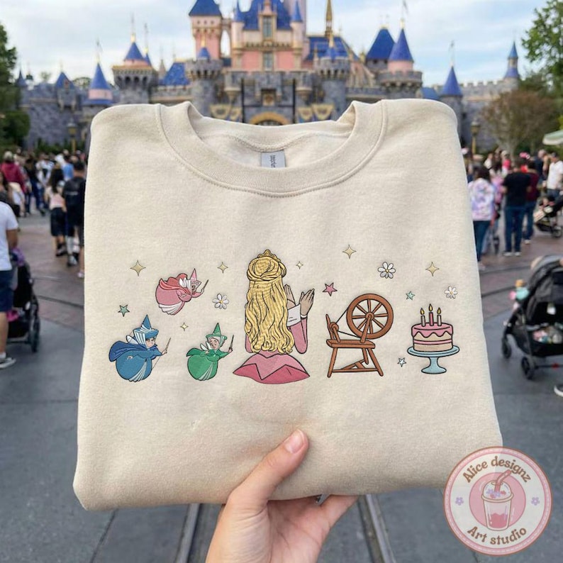 May include: Beige sweatshirt featuring a colorful graphic of a princess, fairies, a spinning wheel, and a cake. The design includes stars and flowers. The sweatshirt is held up in front of a castle.