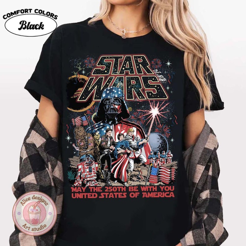 Op de afbeelding: Zwart T-shirt met een Star Wars-afbeelding met Darth Vader in een Amerikaans vlagontwerp. Het shirt bevat de tekst "MAY THE 250TH BE WITH YOU UNITED STATES OF AMERICA". Het shirt heeft ook de woorden "COMFORT COLORS" en "Black" erop gedrukt.