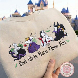 May include: Beige sweatshirt featuring embroidered Disney villains: Ursula, Evil Queen, Cruella de Vil, and Maleficent. The phrase "Bad Girls Have More Fun" is embroidered below the characters, with bats and green accents.
