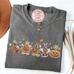 Puede incluir: Una camiseta gris carb&oacute;n con una fila de personajes de dibujos animados disfrazados de calabazas. Los personajes incluyen a Goofy, el Pato Donald, Mickey Mouse, Minnie Mouse y Pluto. La camiseta tiene una pegatina redonda de Alice Designz Art Studio.
