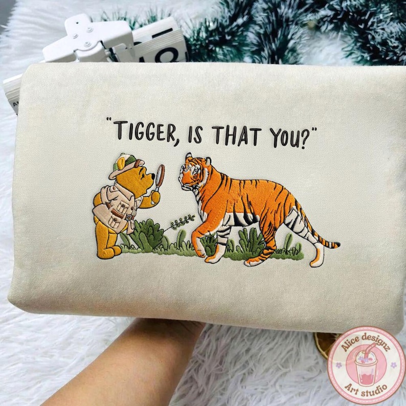 Op de afbeelding: Een beige rechthoekig kussen met een geborduurd ontwerp met Winnie de Poeh en Tijgertje. De tekst "Tigger, is that you?" staat boven de personages. Het ontwerp bevat groen gras en een vergrootglas.