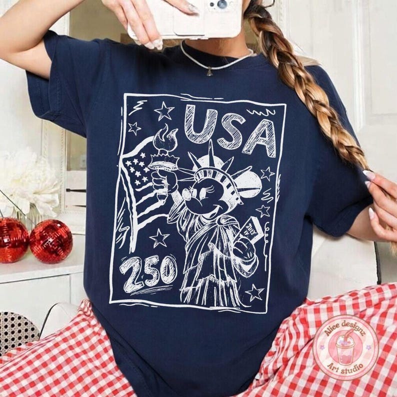 May include: Navy blue t-shirt with a white graphic featuring the Statue of Liberty as a cartoon character, holding a torch and a scroll. The word "USA" is above the character, with the number "250" below. Stars and a waving American flag complete the design.
