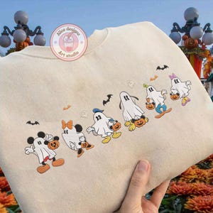 K&ouml;nnte beinhalten: Beiges Sweatshirt mit gestickten Cartoon-Figuren, die als Geister verkleidet sind und jeweils einen K&uuml;rbis halten. Zu den Figuren geh&ouml;ren Micky und Minnie Maus sowie andere Disney-Figuren. Flederm&auml;use sind &uuml;ber den Figuren gestickt. Das Sweatshirt tr&auml;gt den Text "Alice designz Art studio".