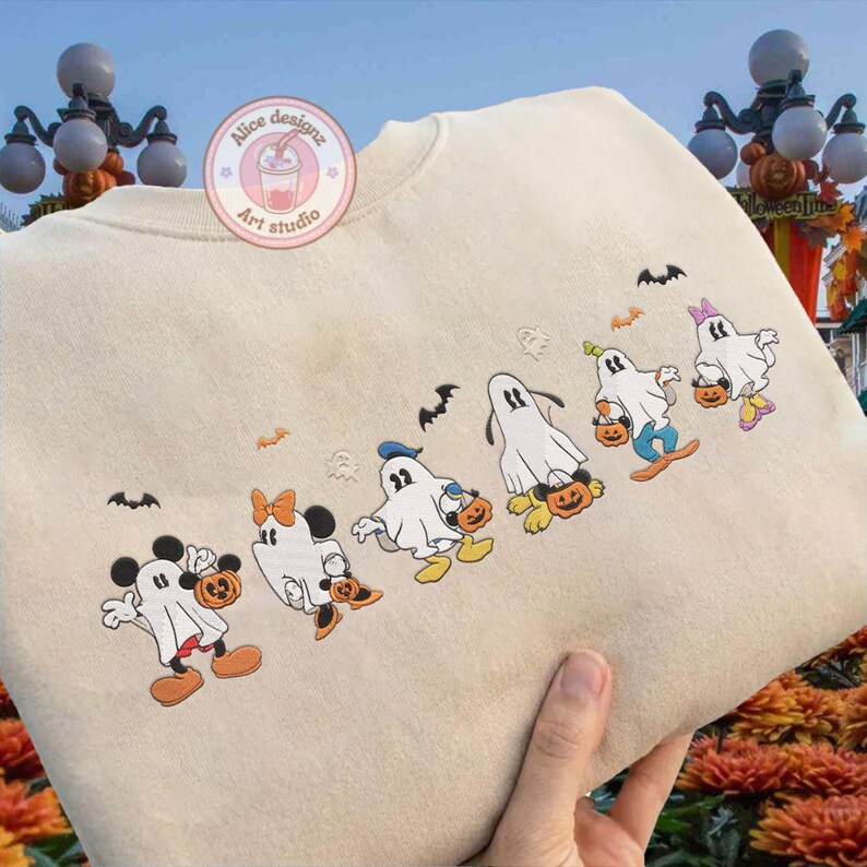 K&ouml;nnte beinhalten: Beiges Sweatshirt mit gestickten Cartoon-Figuren, die als Geister verkleidet sind und jeweils einen K&uuml;rbis halten. Zu den Figuren geh&ouml;ren Micky und Minnie Maus sowie andere Disney-Figuren. Flederm&auml;use sind &uuml;ber den Figuren gestickt. Das Sweatshirt tr&auml;gt den Text "Alice designz Art studio".