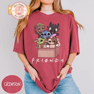 Peut inclure: T-shirt de couleur cramoisie avec un motif de personnages de dessins anim&eacute;s dans une bo&icirc;te et le mot "FRIENDS" en dessous. Le t-shirt a une coupe ample et des manches courtes. Le motif comprend des personnages comme Groot, Stitch et Krokmou.
